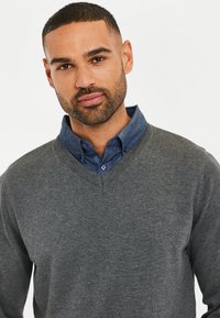 Maglione grigio lavorato a maglia con scollo a V, indossato sopra una camicia blu con bottoni. Texture liscia, vestibilità casual, con un design del colletto a strati.