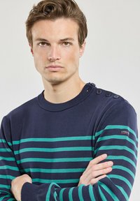 Pull bleu marine avec des rayures horizontales vertes, tissu texturé, poignets et col côtelés, avec quatre boutons décoratifs sur l'épaule.