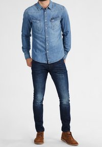 Chemise en denim bleu clair à manches longues, avec poches poitrine et coutures contrastantes, associée à un jean sombre et des chaussures en suède marron.