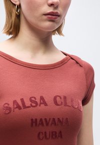 Νεαρός με κοντά ξανθά μαλλιά φοράει ένα πουκάμισο χρώματος σκουριάς με κεντημένο το "Salsa Club Havana Cuba" και χρυσούς κρίκους στα αυτιά.