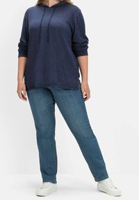 Navyblaues Kapuzensweatshirt mit langen Ärmeln, lockerer Passform und abgerundetem Saum, kombiniert mit hellblauen gerade geschnittenen Jeans und weißen Sneakers.