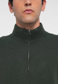 Uomo che indossa un maglione di maglia verde scuro con un colletto alto e una mezza cerniera argento al collo.