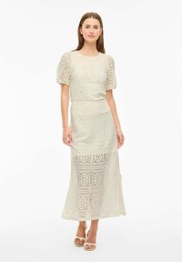 Robe maxi en dentelle tricotée couleur crème avec manches courtes bouffantes et motif floral. Texture lisse avec une taille cintrée et une jupe longue.