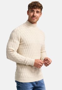 Suéter de cuello alto tejido en color crema con un patrón de olas texturizado, puños y dobladillo acanalados, combinado con unos pantalones de mezclilla azul.