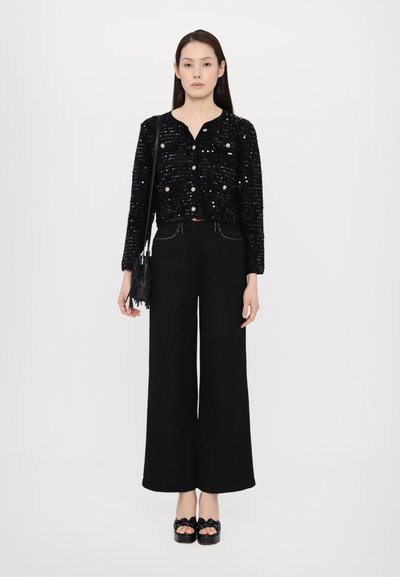Cardigan scurt negru cu paiete, purtat cu pantaloni largi negri cu cusături albe. Modelul ține o geantă mică neagră cu franjuri. Pantofii cu toc negri completează ținuta.