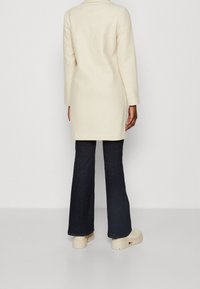 Crème wollen blend jas met een klassiek ontwerp, knielang, met lange mouwen en zijsplitten, gecombineerd met donkere flare jeans en beige plateau schoenen.