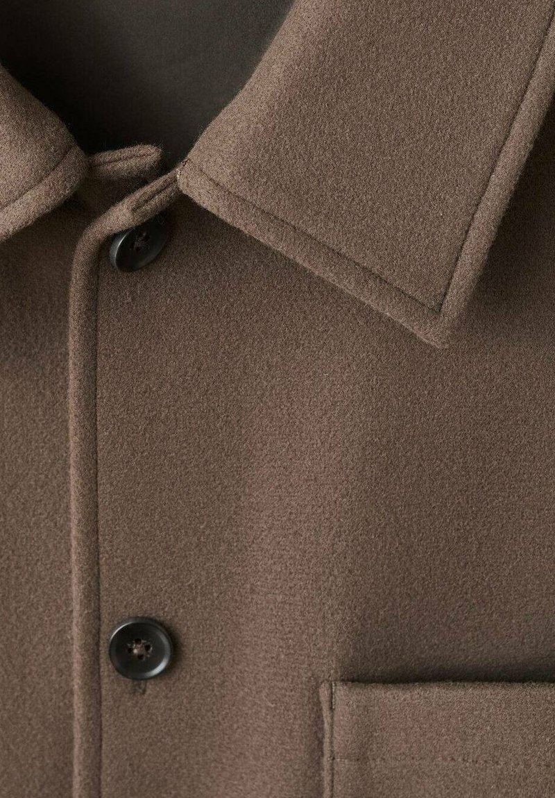 Cappotto di lana marrone con ampio colletto, due bottoni neri e una tasca visibile sul lato destro inferiore.