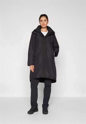 Abrigo negro largo e impermeable con capucha, cremallera frontal y cierres a presión, usado sobre pantalones oscuros y zapatos. Textura suave, diseño minimalista.