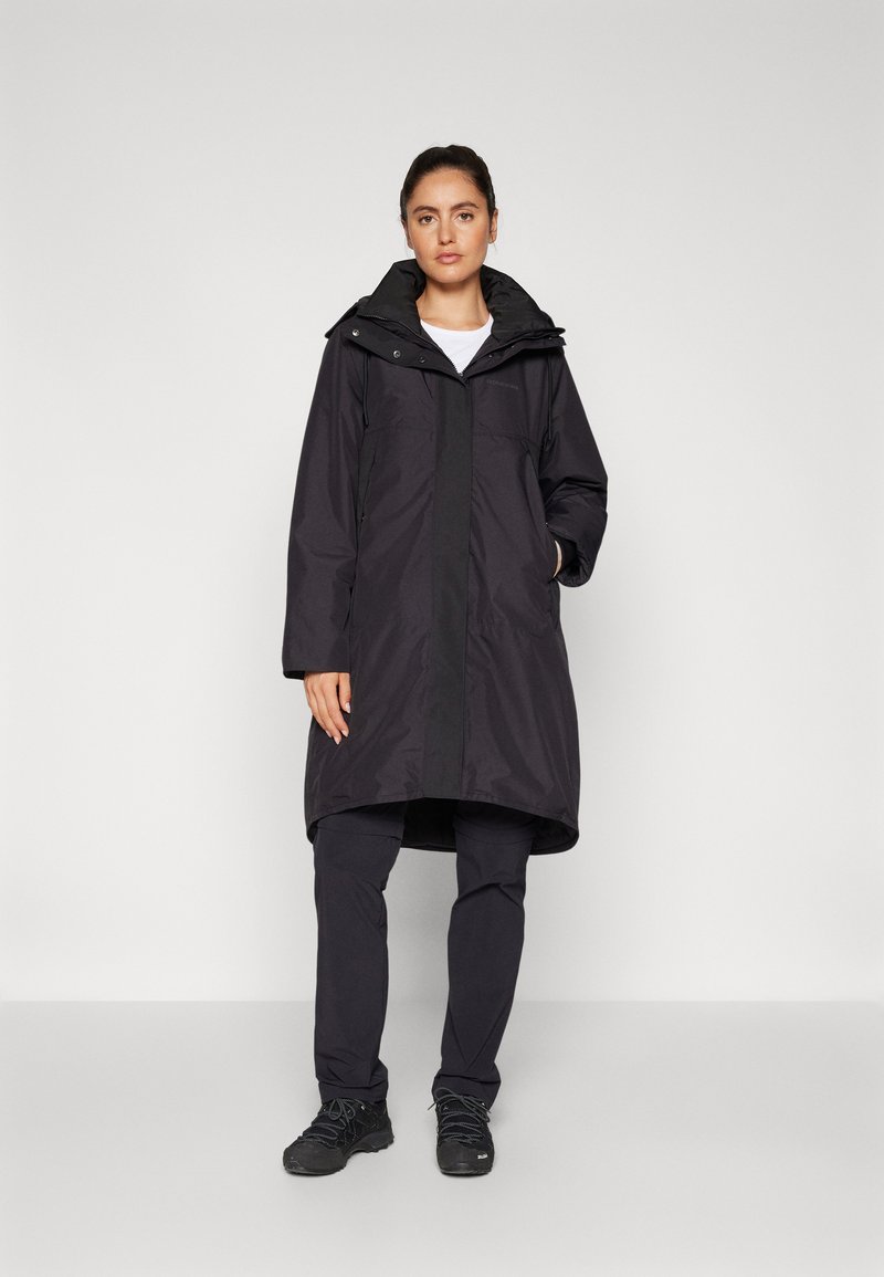 Cappotto lungo nero impermeabile con cappuccio, zip frontale e chiusure a scatto, indossato sopra pantaloni scuri e scarpe. Texture liscia, design minimale.