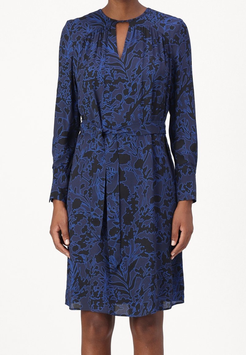 Femme portant une robe bleu marine avec manches longues, un lien à la taille à l'avant et un motif floral noir.