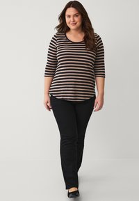 Gestreiftes, langärmliges Oberteil in Marineblau und Beige, kombiniert mit schwarzen Jeans. Das Oberteil hat einen runden Ausschnitt und einen Saum mit Seitenschlitzen. Flache schwarze Schuhe.