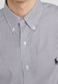 Blå och vit randig skjorta med krage och korta ärmar, med två knappstängningar och en liten svart logotyp på bröstet.