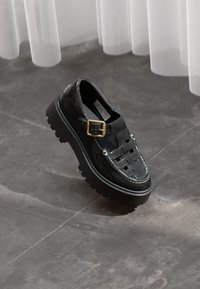 Sapatos de verniz preto com um design trançado e uma fivela dourada, apresentando uma sola chunky e detalhes de costura branca em contraste.