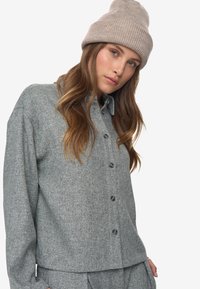 Woman with long hair wears beige knit beanie and matching gray textured button-up shirt and pants, posing against white background.