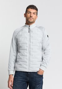 Graue, gesteppte Jacke mit hohem Kragen und Reißverschluss. Verfügt über Raglanärmel und gerippte Bündchen. Kombiniert mit dunklen Jeans.