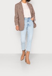 Blazer a cuadros marrón y crema, blusa rosa clara, jeans de mezclilla rotos azul claro y botas de tobillo de tacón beige. Se encuentra de pie contra un fondo neutro.