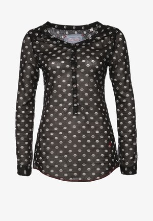 Zwarte doorschijnende blouse met witte stippen, lange mouwen en knoopdetails, gecombineerd met donkere uitlopende spijkerbroeken en glanzend zwarte schoenen.