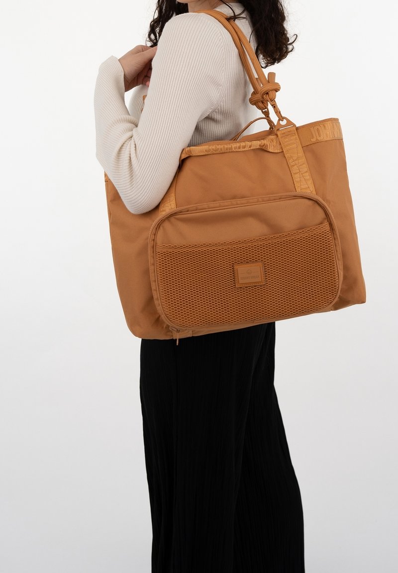 Femme portant un haut beige à manches longues et un pantalon noir tient un grand sac cabas marron avec une poche filet à l'avant sur son épaule.