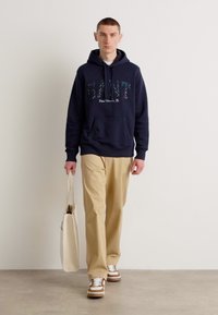 Felpa navy con logo "GANT" in un carattere decorativo, abbinata a pantaloni beige e sneakers marroni e bianchi. Il modello tiene in mano una borsa tote beige.
