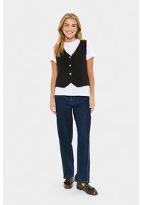 Jonge vrouw in een zwart knoopvest over een wit T-shirt, donkere spijkerbroek en bruine instappers, lachend met ontspannen handen langs haar zij.