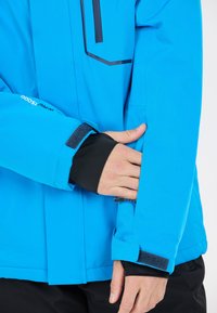 Ljusblå vattentät jacka med svarta tygdetaljer. Har elastiska ärmslut, en ficka med dragkedja och en snygg, modern design.