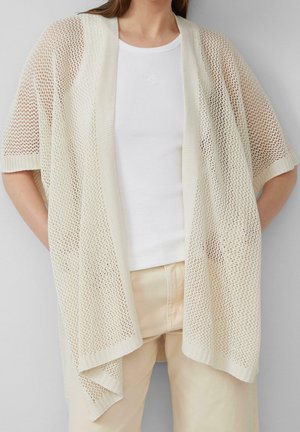 Femme portant un cardigan à manches courtes en maille ouverte beige sur un débardeur côtelé blanc et un pantalon beige clair, debout avec les mains dans les poches.