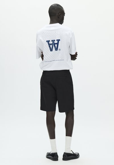 T-shirt en coton blanc avec un logo graphique bleu dans le dos, associé à un short noir. Le mannequin porte des mocassins noirs et des chaussettes blanches.