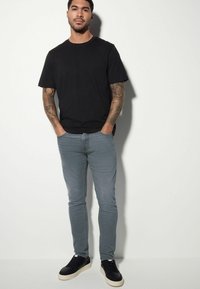 Tricou negru din bumbac, blugi gri slim-fit și adidași negri cu sole albe. Bărbatul are tatuaje vizibile pe ambele brațe.