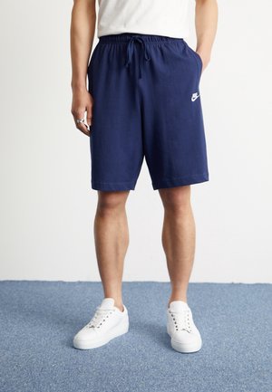 Man draagt marineblauwe Nike-short met trekkoord en witte sneakers, staande op een blauw tapijt.