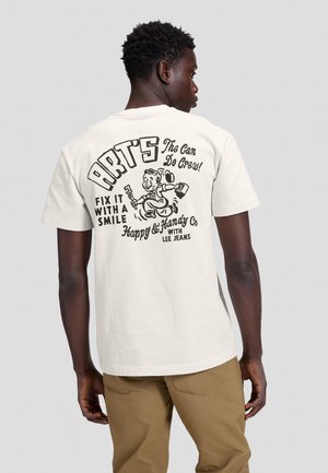 Homme en pantalon beige portant un T-shirt blanc avec un bricoleur en dessin animé et le texte ""Art’s fix it with a smile"" dans le dos.