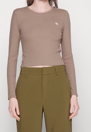 Femme portant un haut beige ajusté à manches longues avec un petit logo d'élan blanc et un pantalon taille haute vert olive.