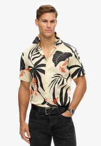 Camisa de manga corta con fondo crema, que presenta grandes patrones de hojas tropicales en negro y naranja y un diseño de cuello abotonado.