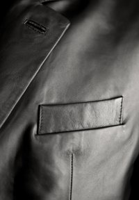 Szczegół czarnej skórzanej kurtki z gładką fakturą, klapką na kołnierzu oraz prostokątną kieszenią po lewej stronie z widocznym ściegem.