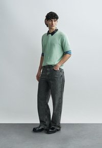 Gaiši zaļš teksturēts džemperis ar īsām piedurknēm, valkāts pār tumšu krekla apkakli, kombinēts ar vaļējām pelēkām džinsām un melniem apaviem.