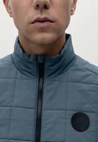 Giacca trapuntata di colore blu scuro con colletto alto e zip nera. Presenta un logo rotondo sul petto. Tessuto morbido e design sobrio.