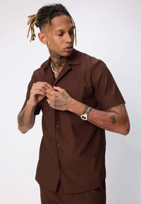 Chemise à manches courtes brune avec un fini texturé et un col, portée par une personne qui ajuste les boutons, avec des tatouages visibles.