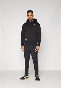 Black hoodie with a front pocket and gold logo, paired with black pants and black and tan shoes. Smooth fabric, relaxed fit.