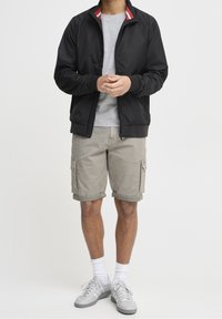Veste bomber noire avec col rayé rouge et blanc, sweat-shirt gris, shorts cargo et baskets grises. Modèle debout, mains jointes.