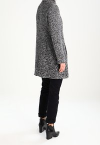 Grå og hvidt mønstret frakke med høj krave, knælængde, båret over sorte bukser og ankelstøvler, med en struktureret, tekstureret stof.