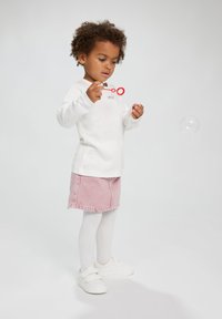 Witte lange mouwen shirt met de tekst "smile", roze spijkerrok, witte panty en witte sneakers; kind dat speelt met een rode bellenblaasstok.