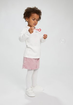 Witte lange mouwen shirt met de tekst "smile", roze spijkerrok, witte panty en witte sneakers; kind dat speelt met een rode bellenblaasstok.