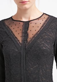 Top de encaje negro con una pechera de malla transparente y lunares negros, mangas largas y un escote en V profundo. Intrincados patrones florales en el encaje.