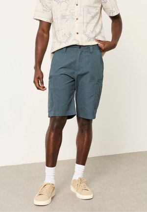 Uomo con pantaloncini blu fino al ginocchio, calzini bianchi, scarpe da ginnastica beige e una maglietta beige a maniche corte con stampe di granchi, in piedi su un pavimento grigio.