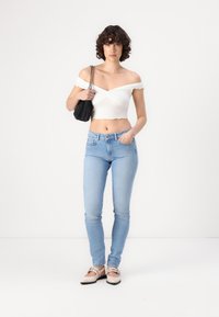 Bela off-shoulder kratka majica s valovitim robom, v kombinaciji s svetlo modrimi oprijetimi kavbojkami in bež balerinkami. Model drži črno torbico.
