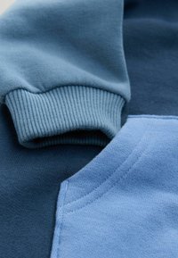 Nahaufnahme von gefalteten blauen Sweatshirtärmeln, die gerippte Bündchen und genähte Taschendetails auf weichem Stoff zeigen.