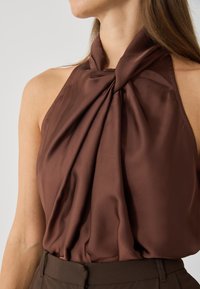 Hnědý halter top vyrobený z hladkého, hedvábného materiálu s rozšířenými detaily kolem výstřihu a ramenní oblasti.