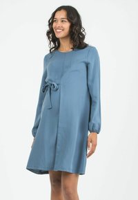 Blaues, knielanges Schwangerschafts-Kleid mit langen Ärmeln, rundem Ausschnitt und einem Schleifen-Detail am Bund, aus weichem Stoff gefertigt.