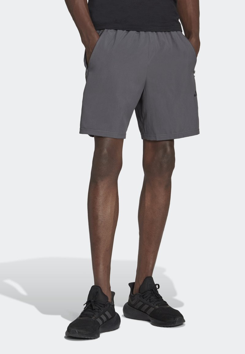 Homme portant un short de sport gris avec poches latérales et des chaussures de course noires, debout les mains dans les poches devant un fond uni.