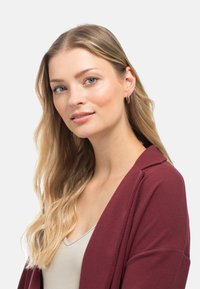 Cardigan borgogna con tessuto testurizzato, frontale aperto e taglio rilassato. Abbinato a una maglietta beige chiaro e piccoli orecchini a cerchio.