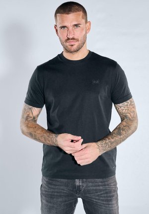 Man with short hair and full sleeve tattoos wears a plain black t-shirt and dark jeans, standing against a light gray background.